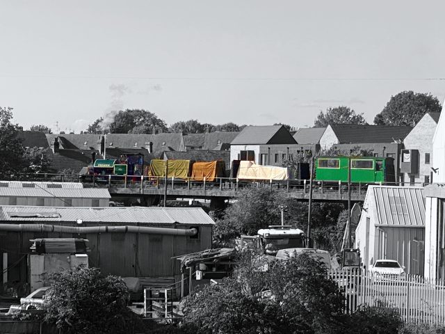 Steam locomotive 'Premier' pulls two wagons and a coach over Milton Regis Viaduct, above the rooftops of the industrial units in the foreground.